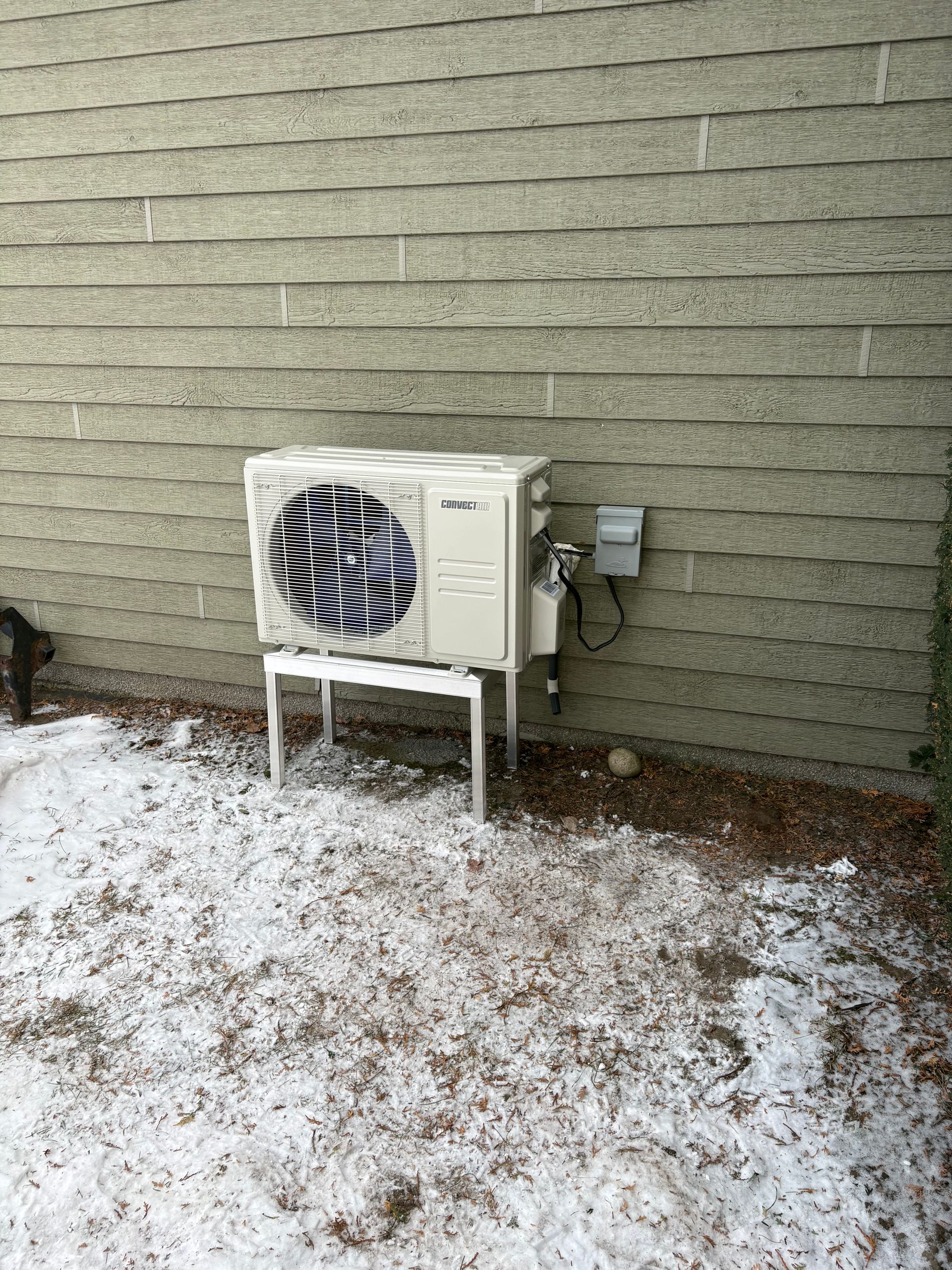 Une unité de pompe à chaleur extérieure mini-split blanc cassé, surélevée sur un support métallique contre un mur de briques, le sol étant enneigé.