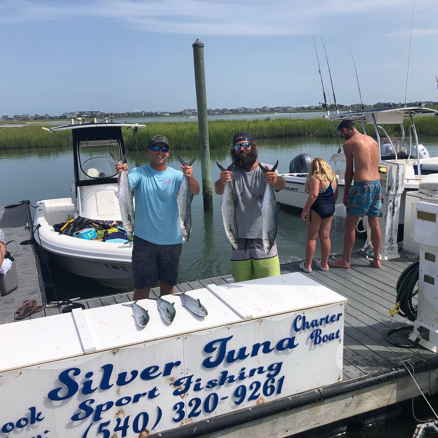 Two men holding fish next to a boat on a dock; others nearby. 