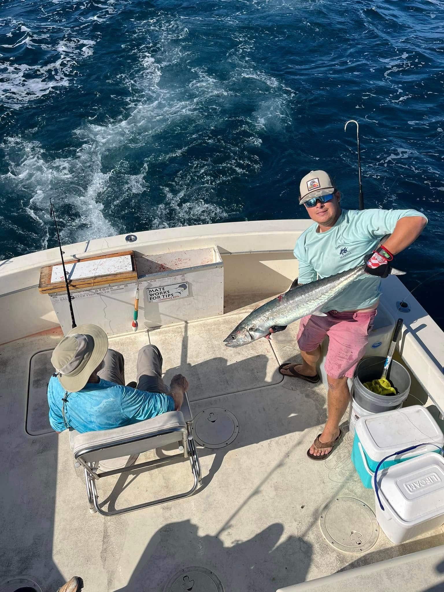 Man on boat holds up fish, another person sits. Ocean in background, sunny day.