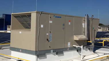 A large, beige Daikin rooftop HVAC unit sits on a flat, white-roofed building against a clear blue sky.