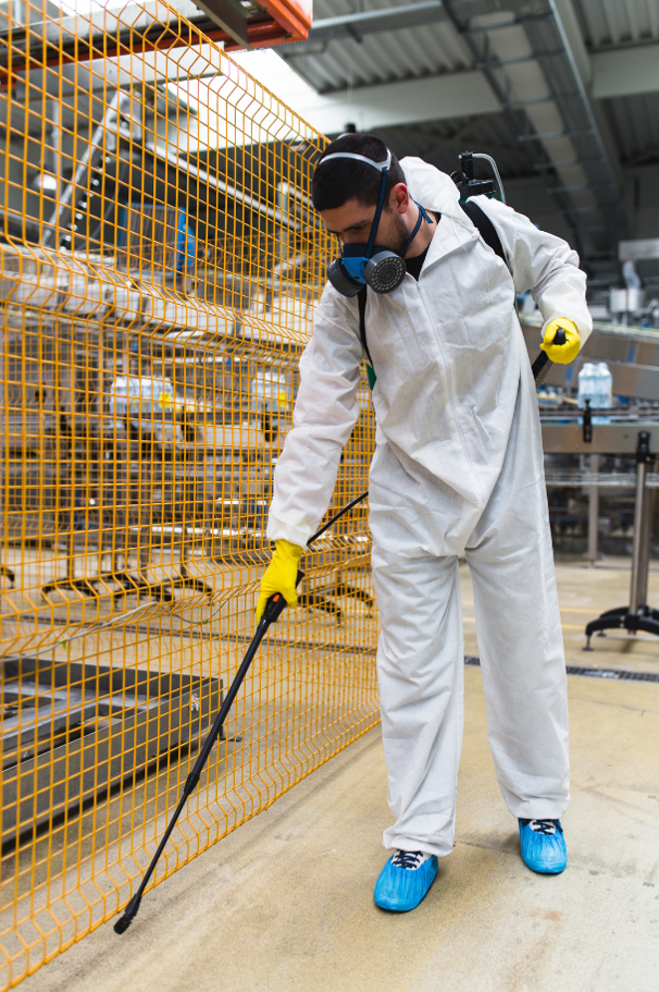 Pest control worker wearing protective suit while spraying pesticides in industrial setting.