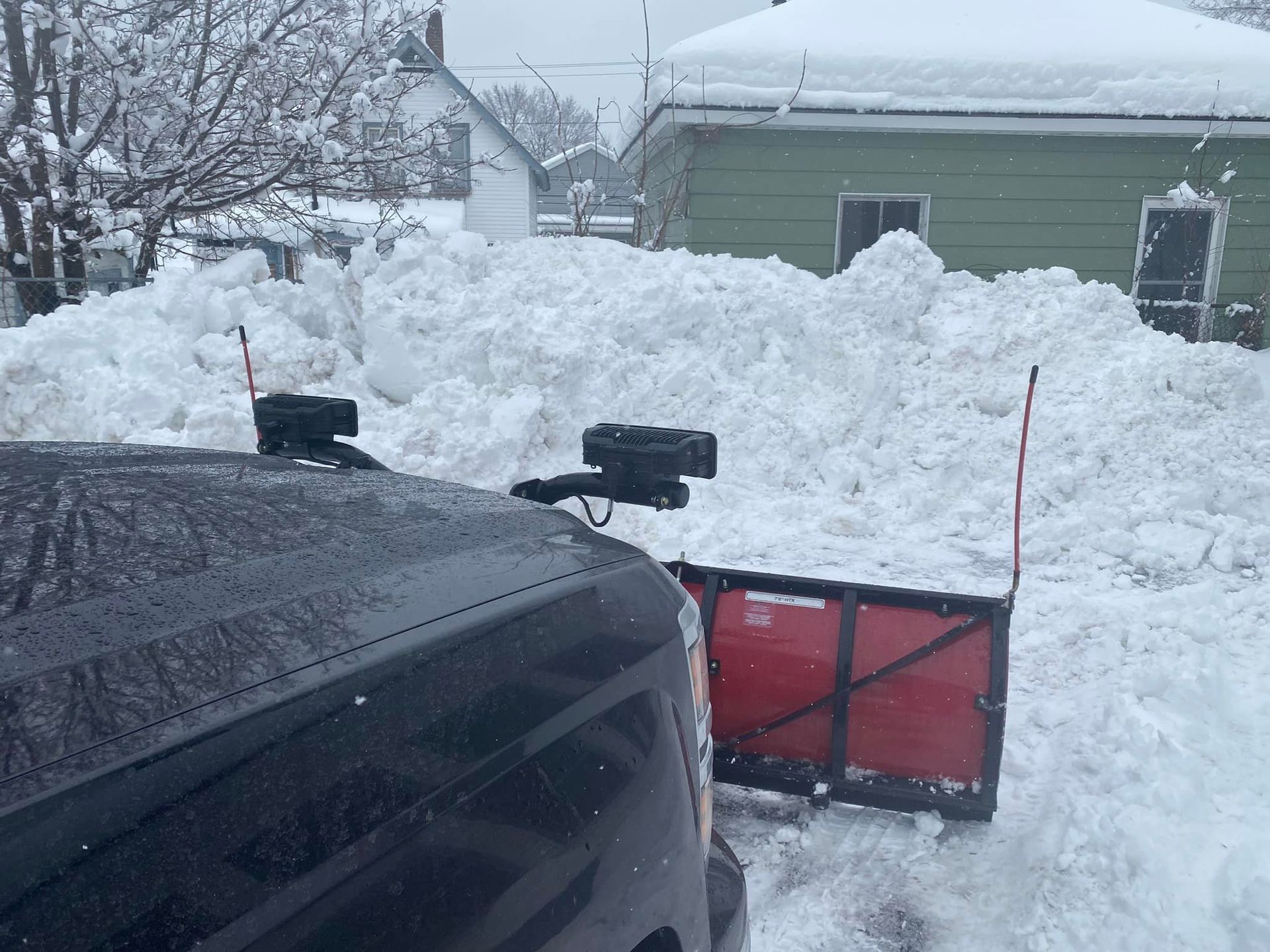 Snow-covered truck with a plow clearing a large snowbank in front of a house.