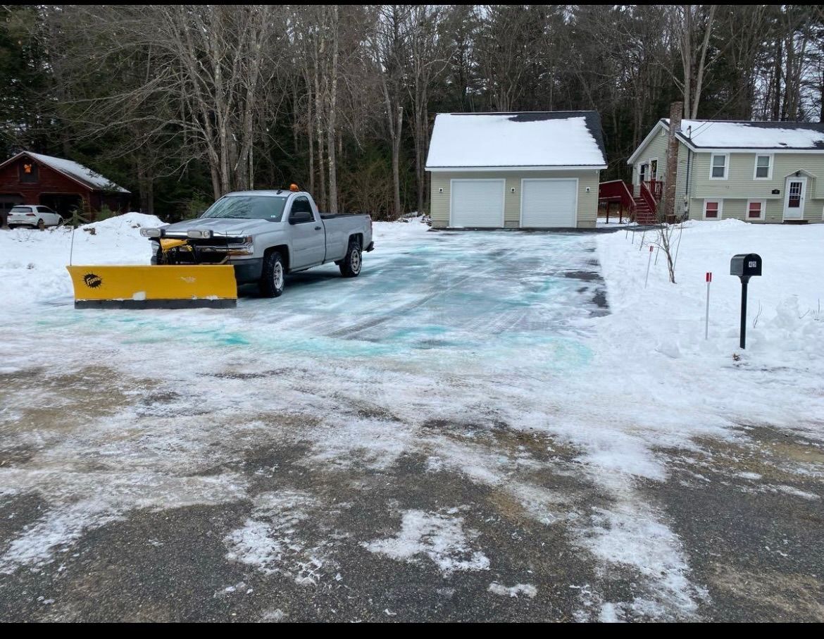 A truck with a snowplow clearing a snow-covered driveway. A house and garage are in the background.