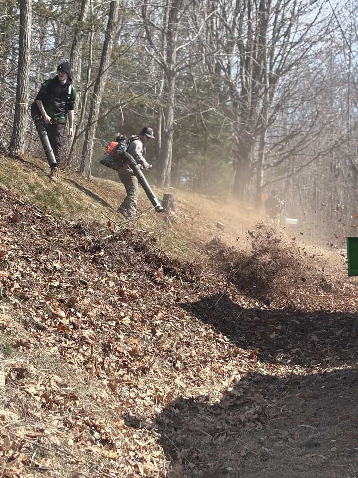 Two people blowing leaves on a hillside trail. Brown leaves and dust are airborne. Trees are in the background.