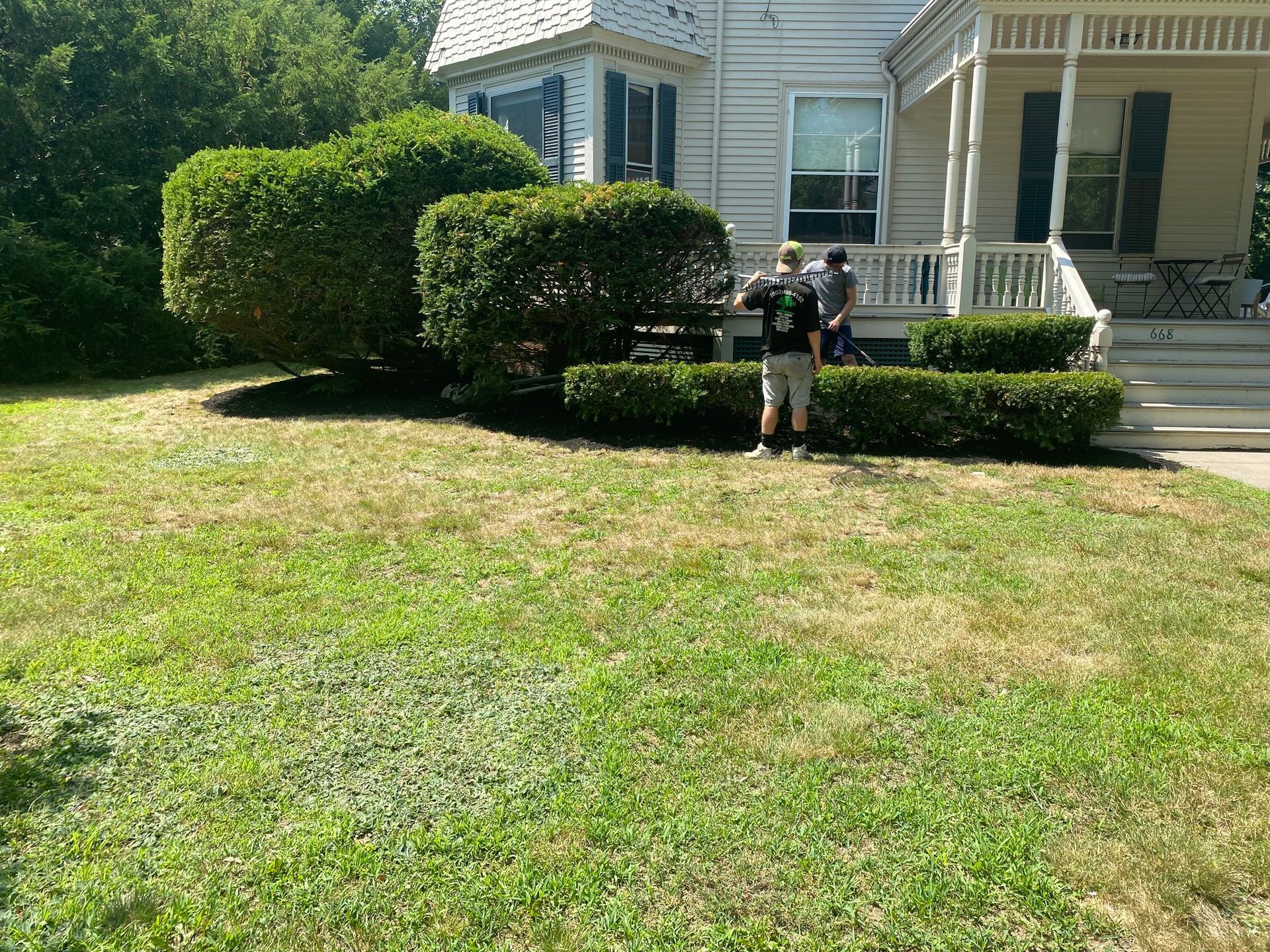 Two people standing near trimmed bushes and a house with porch steps.