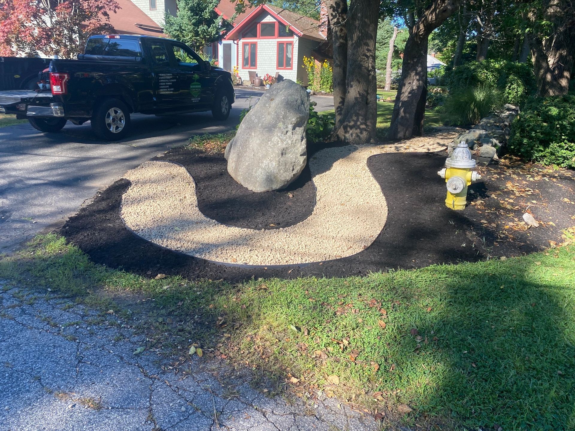 Large boulder in a landscaped garden bed of mulch and gravel beside a driveway; black pickup truck visible.