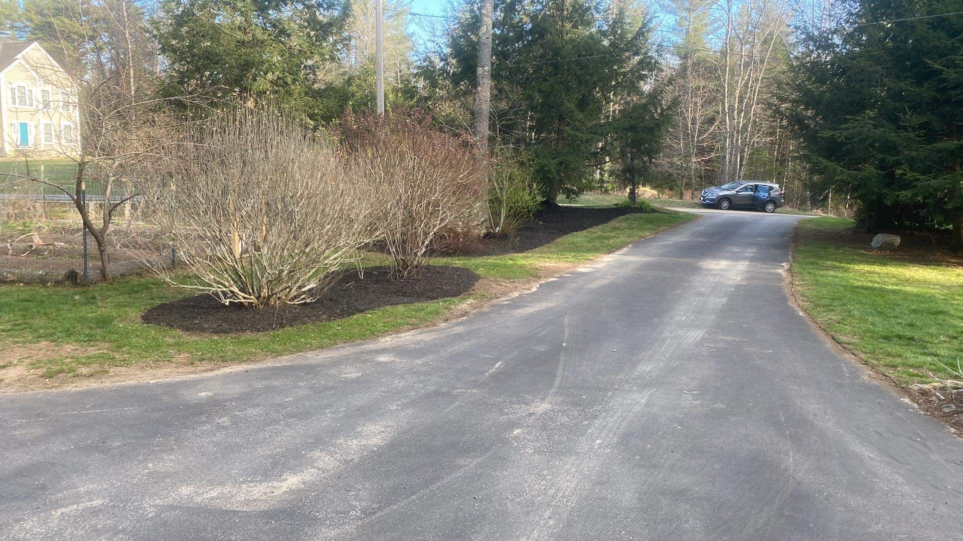 Asphalt driveway leading past mulched landscaping and trees, a car parked at the end.