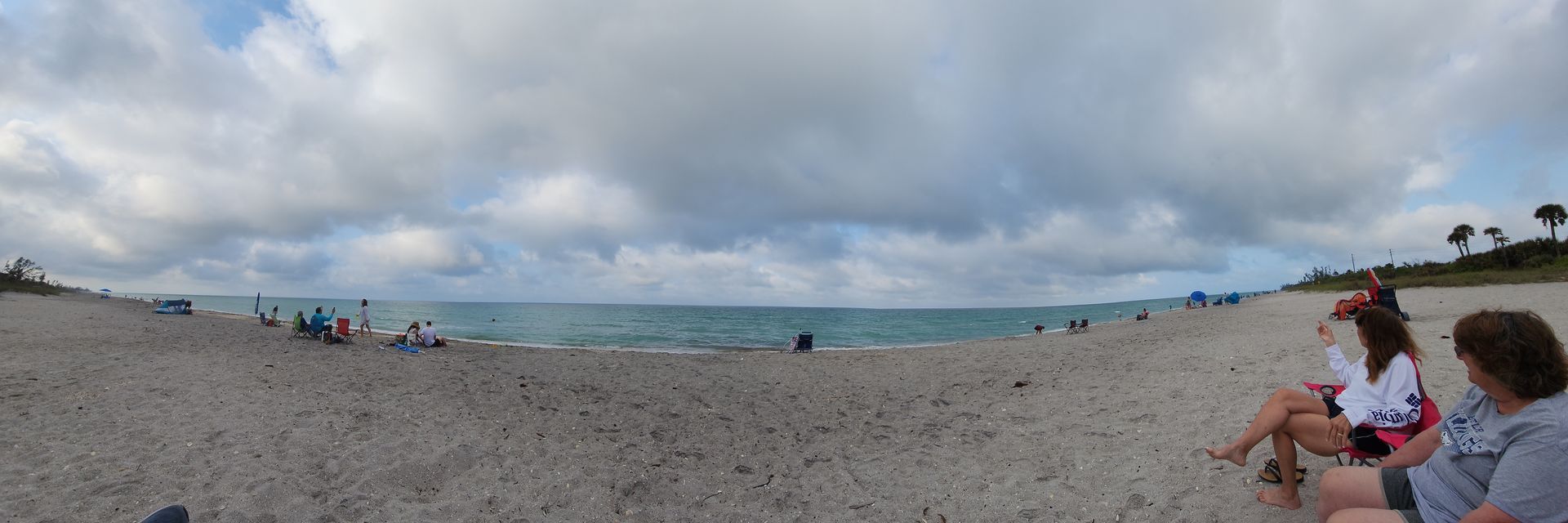 Wide sandy beach under cloudy sky, with people relaxing near the shoreline and ocean in the distance