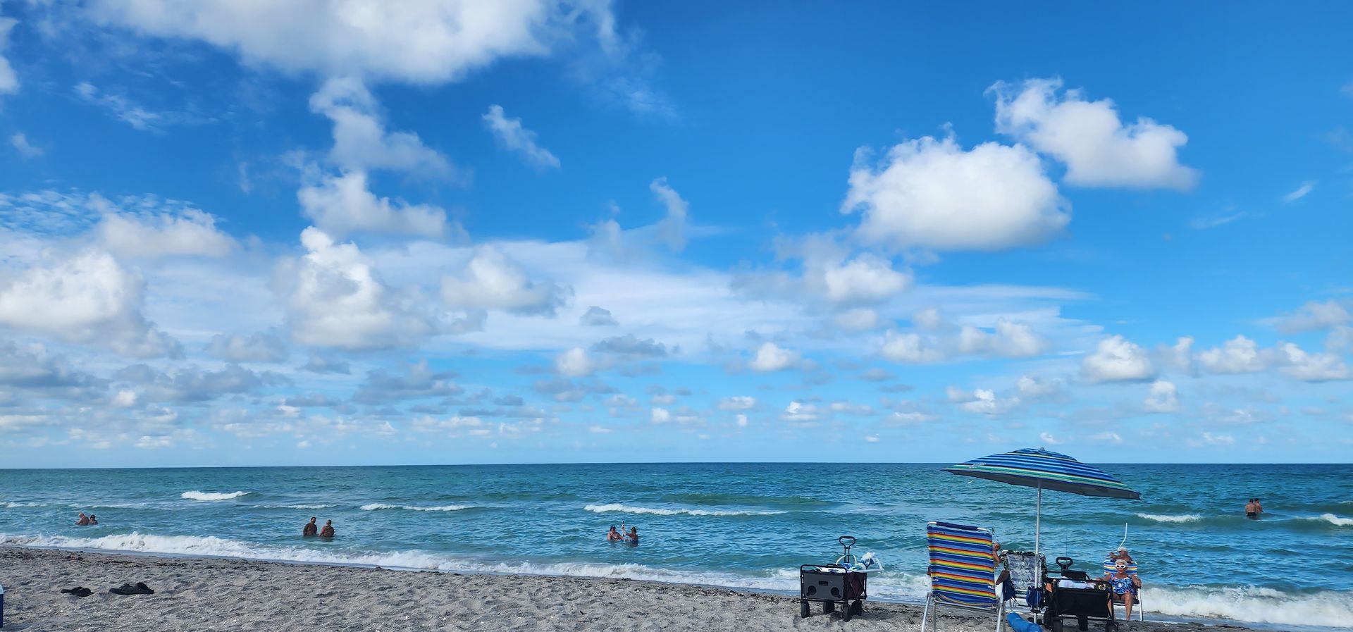 Sunny beach with blue sky, white clouds, ocean waves, and people relaxing on the shore