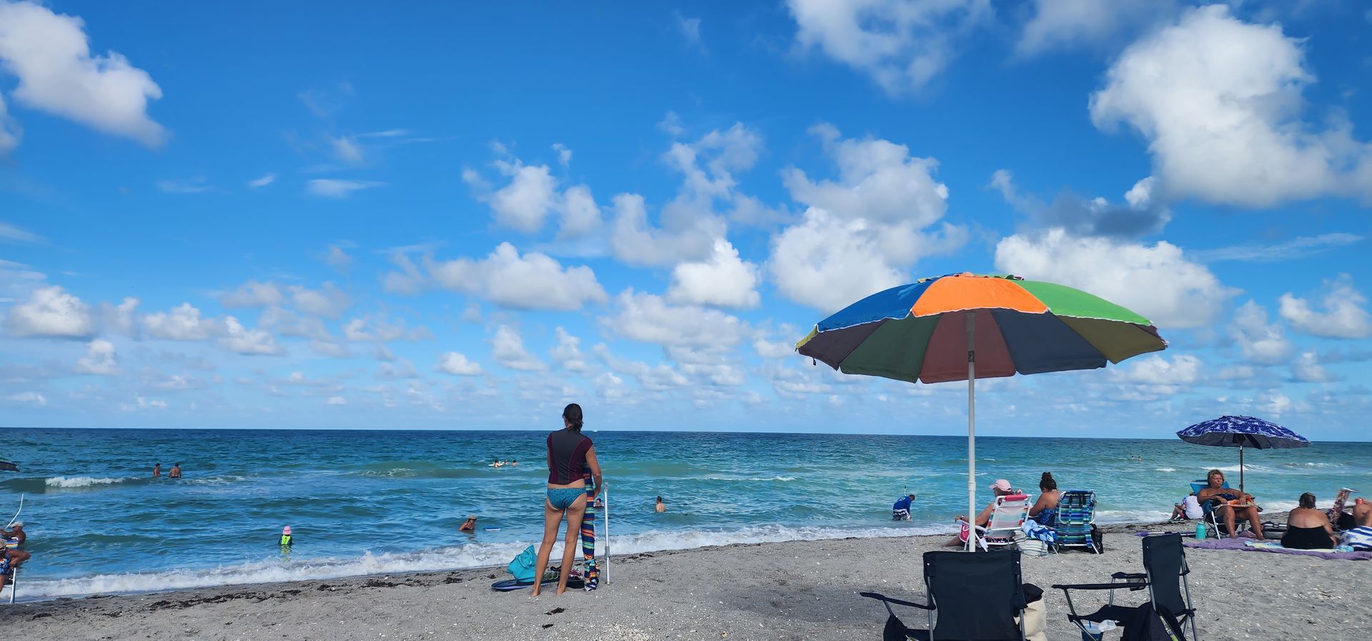 Beach scene with blue ocean, colorful umbrella, and people relaxing under a bright sky