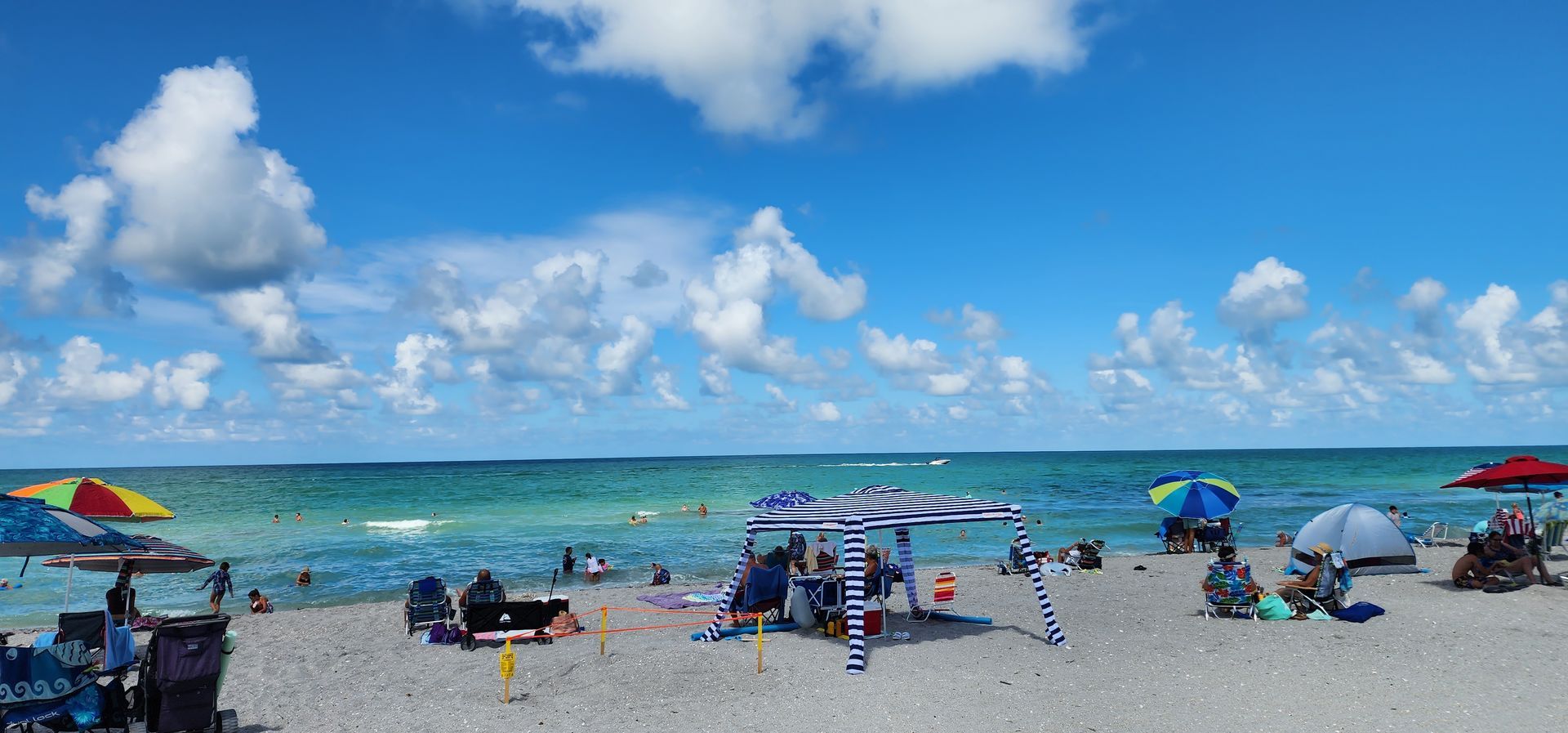 Sunny beach with people under umbrellas, blue ocean, and scattered lounge chairs under a bright sky