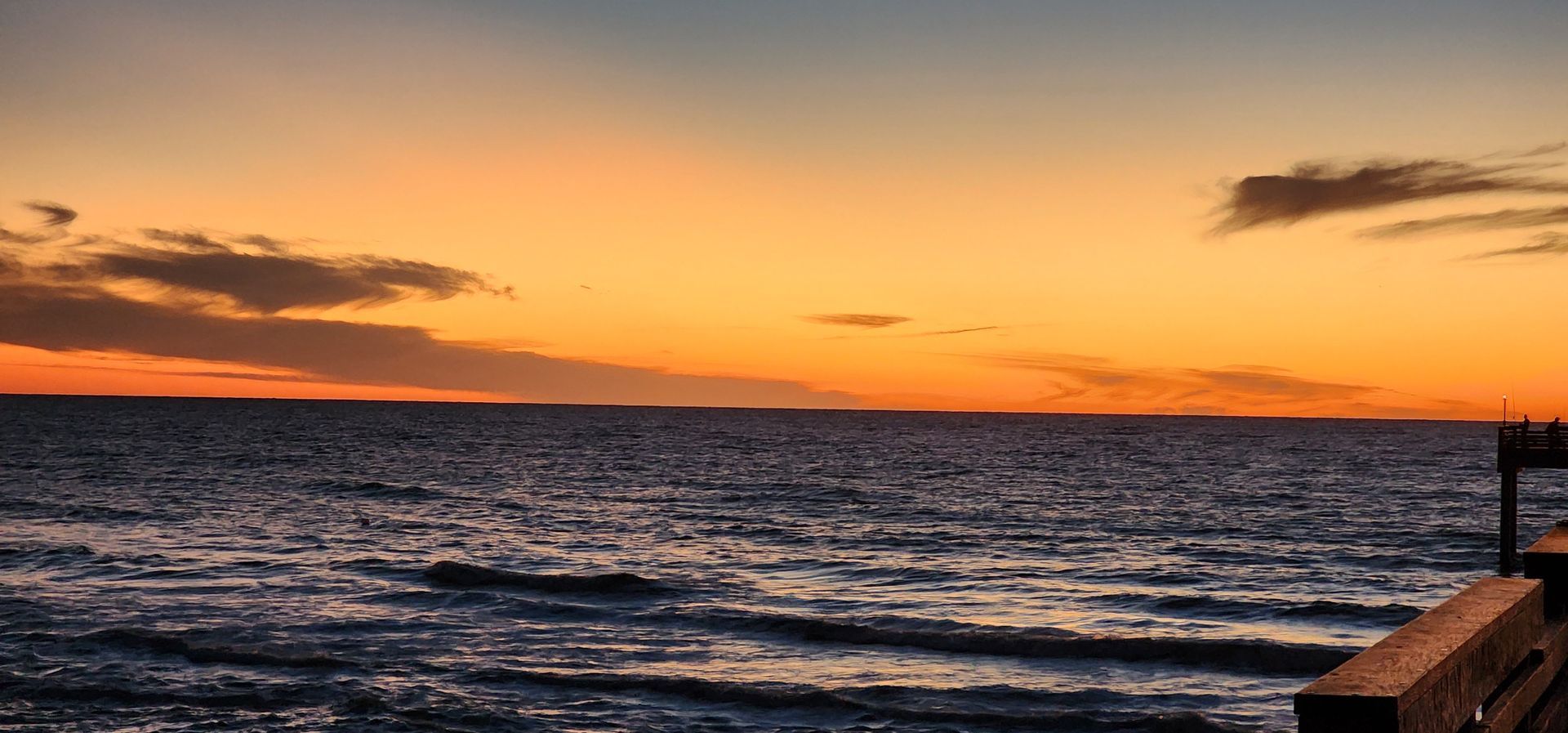 Sunset over the ocean with orange sky, dark clouds, and a wooden pier at right
