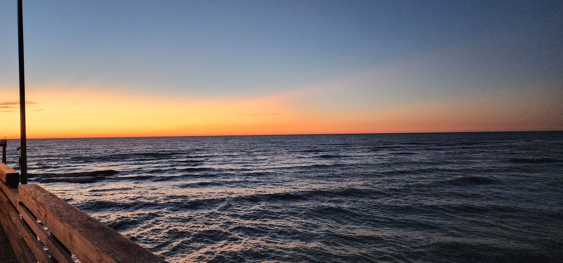 Sunset over a calm ocean, with orange sky and a wooden pier along the left edge