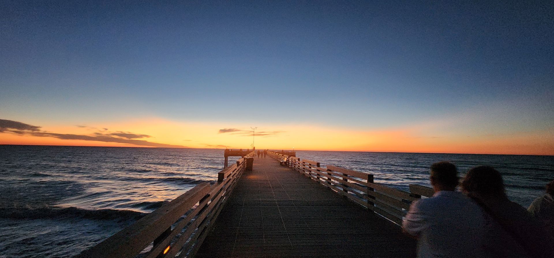 Sunset over a long pier stretching into the ocean, with a person standing at the right edge.