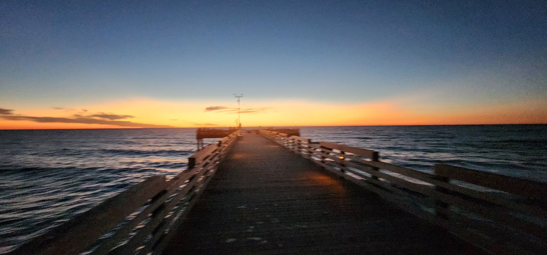 Sunset over a wooden pier extending into calm water with orange sky and dark horizon
