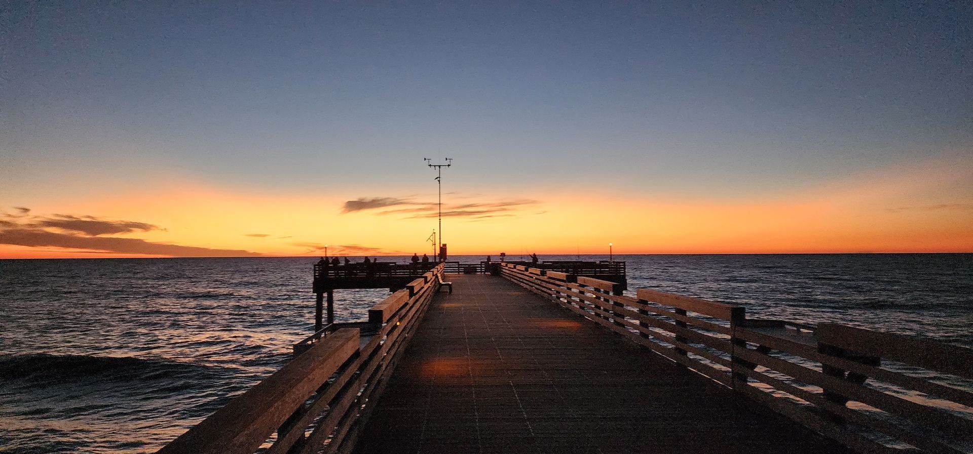 Sunset over a long pier stretching into the ocean, with warm orange sky and calm water.