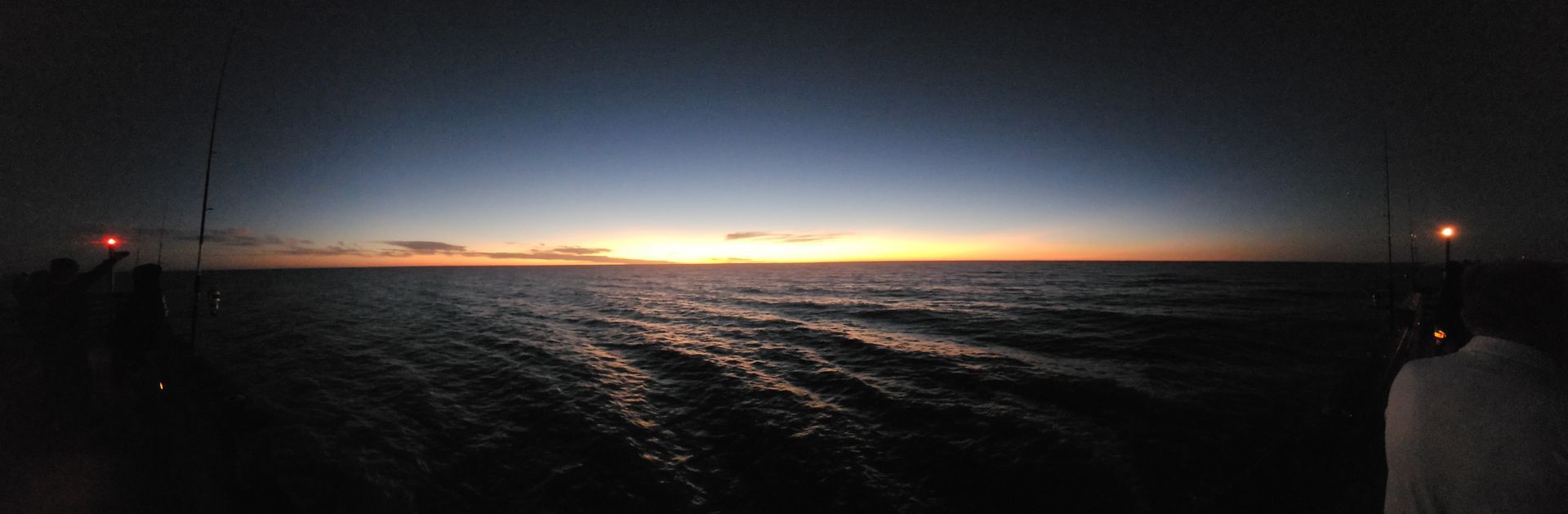 Dark ocean at sunset with a thin orange horizon and distant lights on the water.