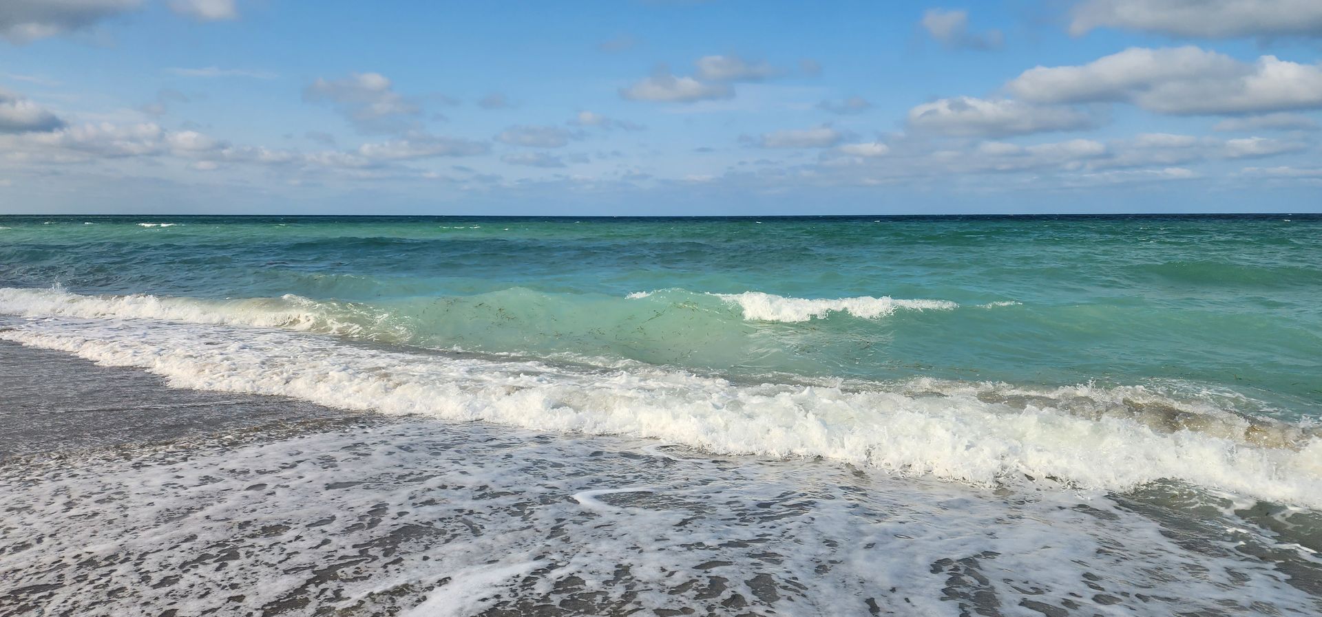 Waves washing onto a rocky beach under a cloudy sky, with turquoise sea and horizon in view
