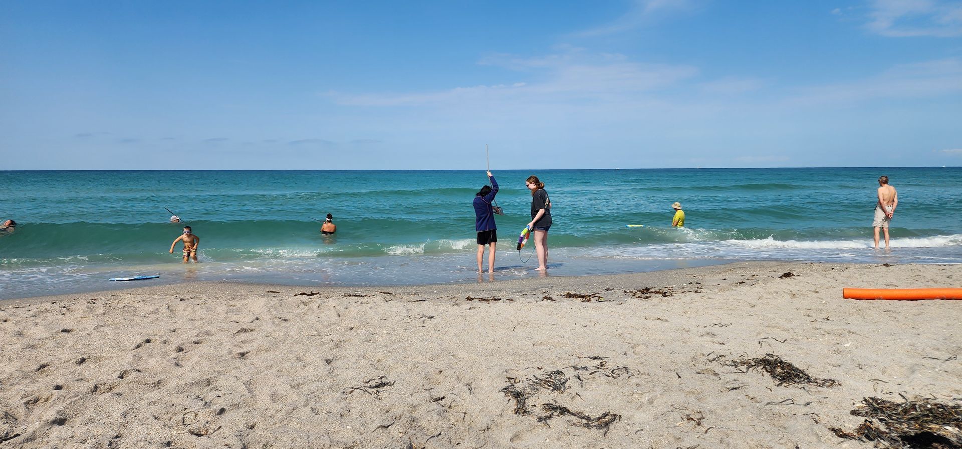 People standing and swimming at a sandy beach with blue ocean and clear sky.