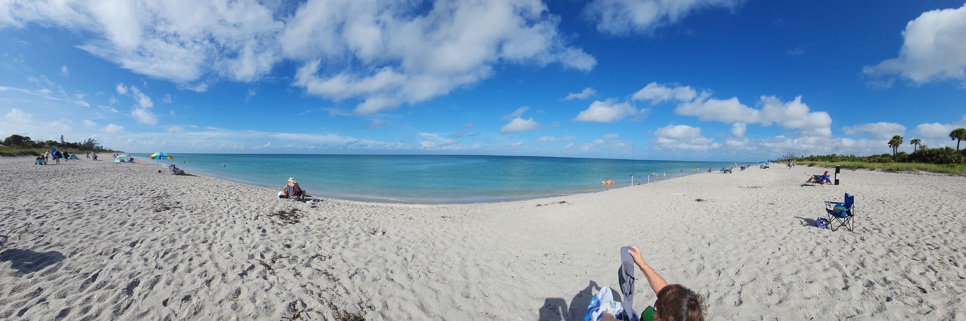 Sunny beach with white sand, blue ocean, and a few people relaxing under a clear sky.