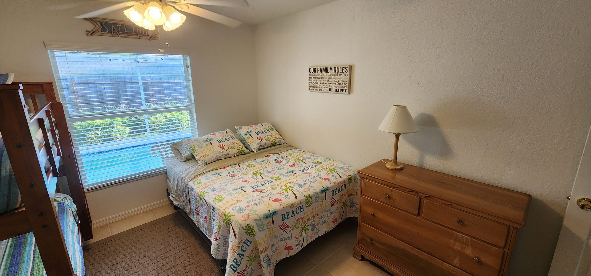 Small bedroom with bed, dresser, lamp, and window with blinds opening to a view outside