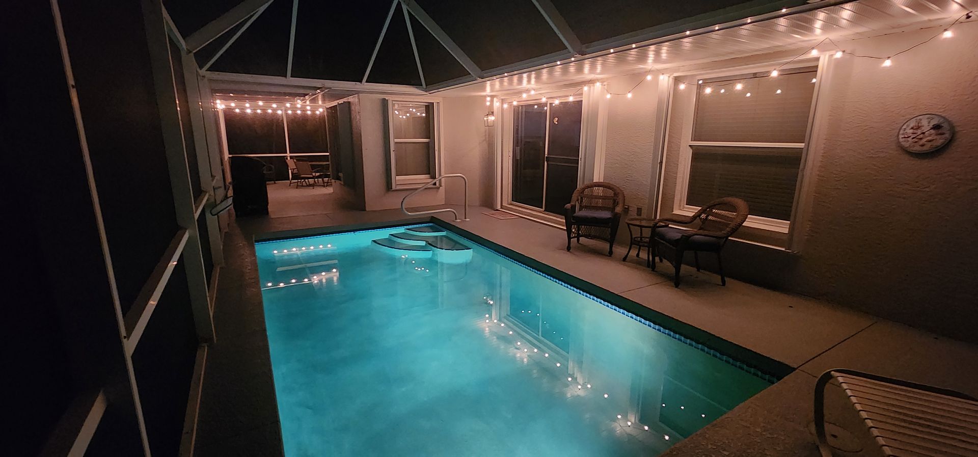A lit backyard pool at night with blue water, lounge chairs, and covered patio seating.