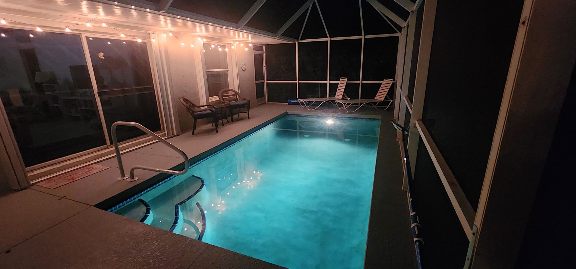 Nighttime pool under a screened patio with turquoise water, deck lights, and lounge chairs