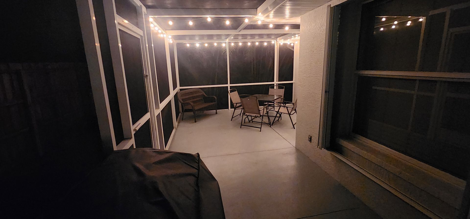 Covered patio with string lights, a table and chairs in the distance, and a grill in the foreground at night