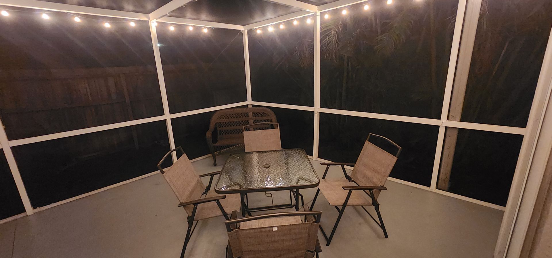 Screened porch with string lights, a glass table, and four folding chairs at night