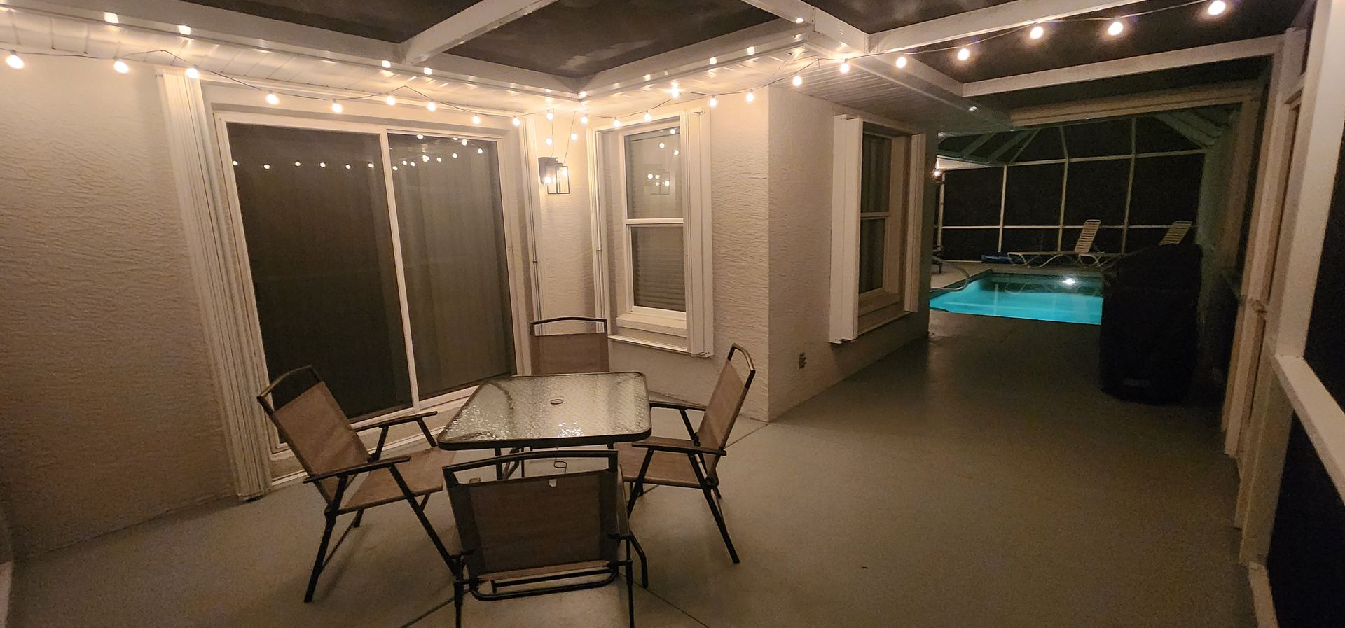 Patio with a glass table and chairs under string lights, opening to a lit pool at night