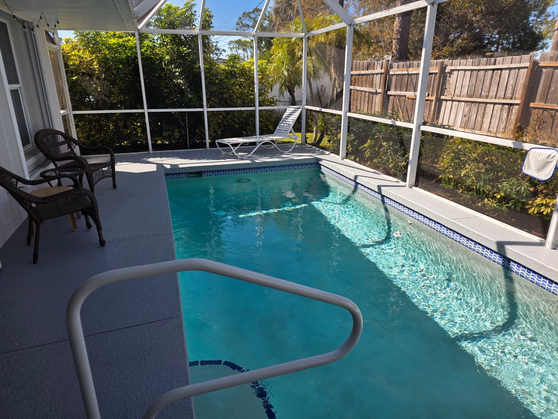 Screened-in backyard pool with turquoise water, patio chairs, and a wooden fence in bright sunlight