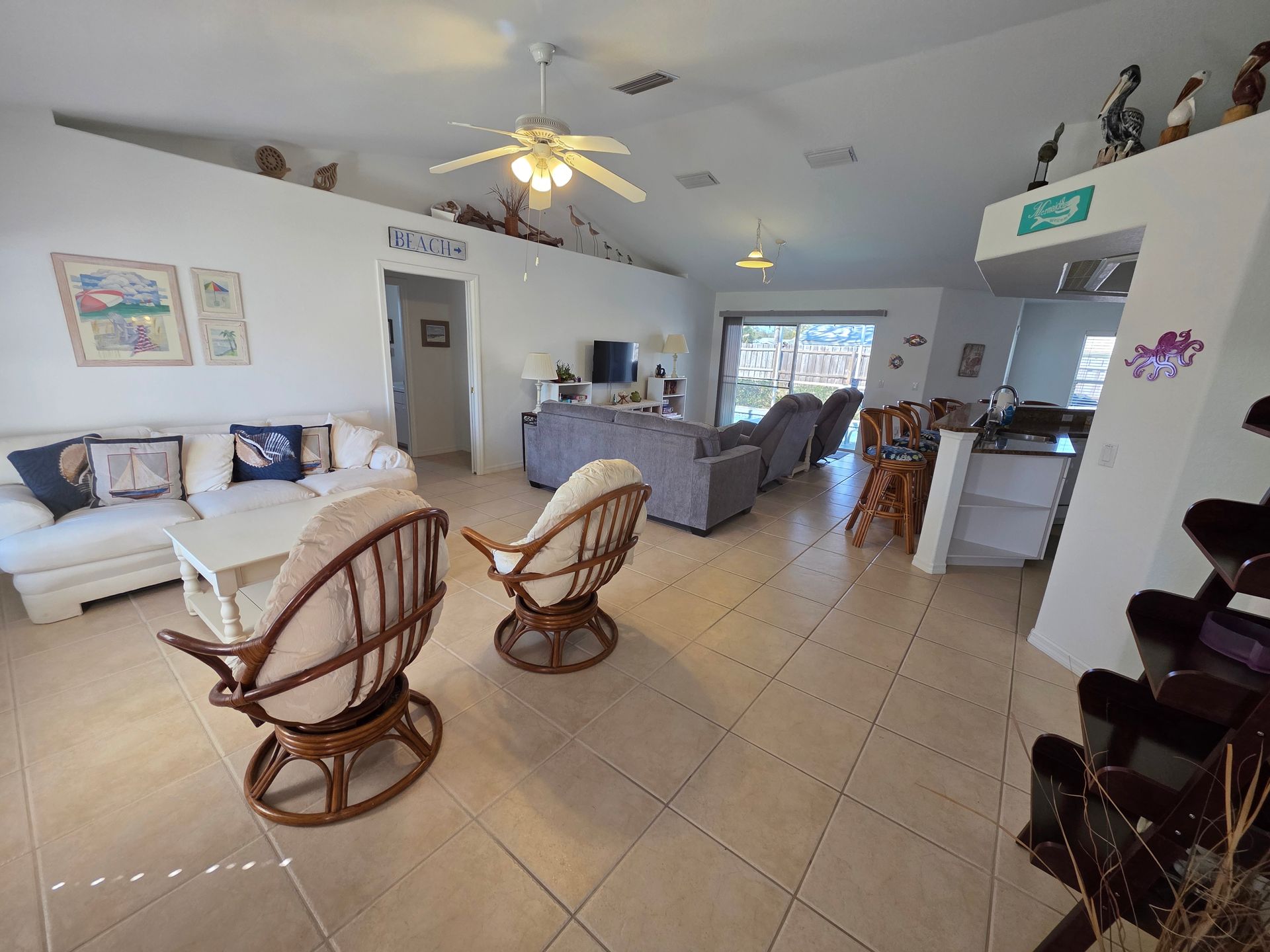 Bright, open living room with white sofa, wicker chairs, tiled floor, and kitchen area in back