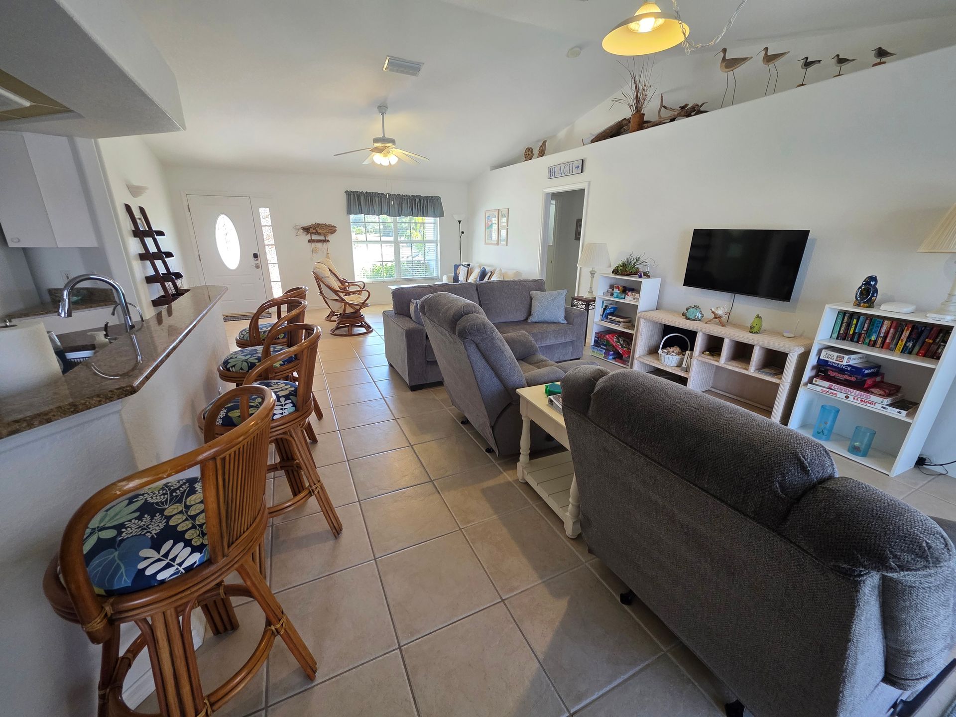 Bright open living room with gray sofas, bar stools, TV, and tiled floor leading to a sunny back door