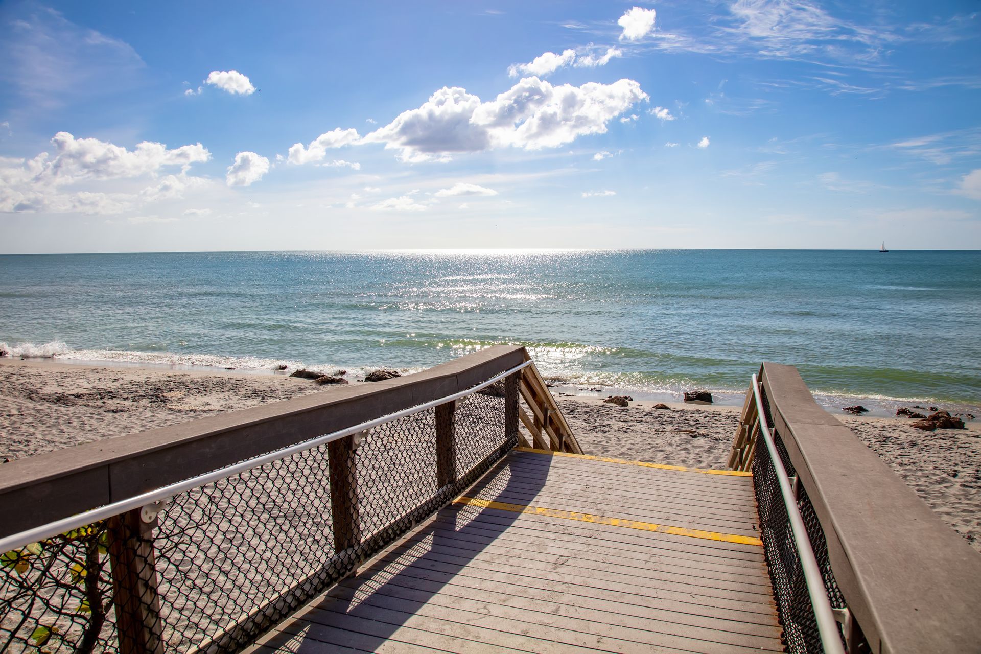 Wooden beach walkway leading to a sunny ocean shoreline under a blue sky with clouds Wooden beach walkway leading to a sunny ocean shoreline under a blue sky with clouds