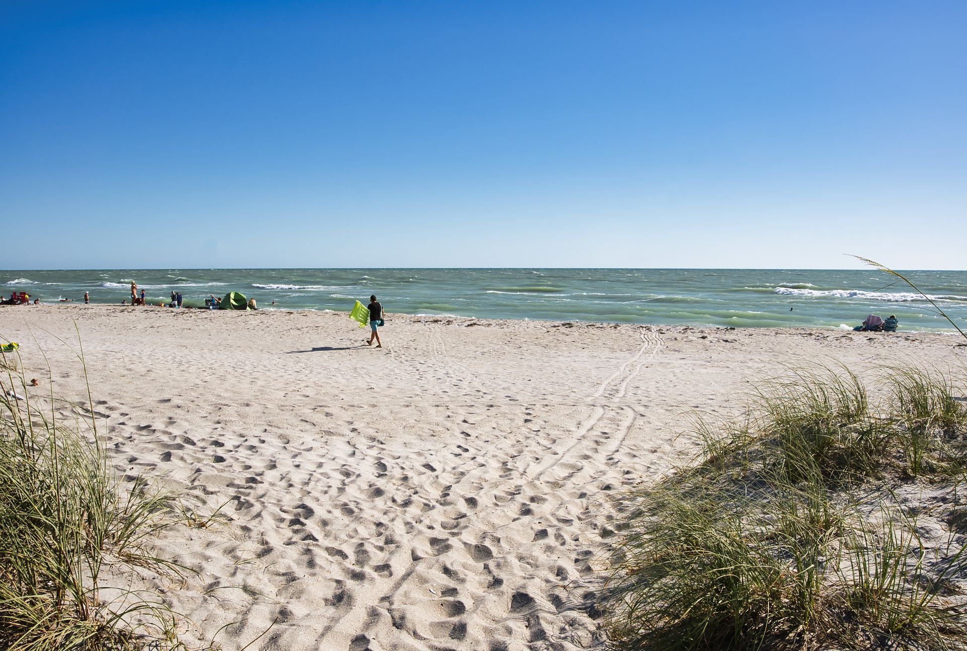 Sandy beach with dunes, a person near the shoreline, and a blue sky over choppy water. Sandy beach with dunes, a person near the shoreline, and a blue sky over choppy water.