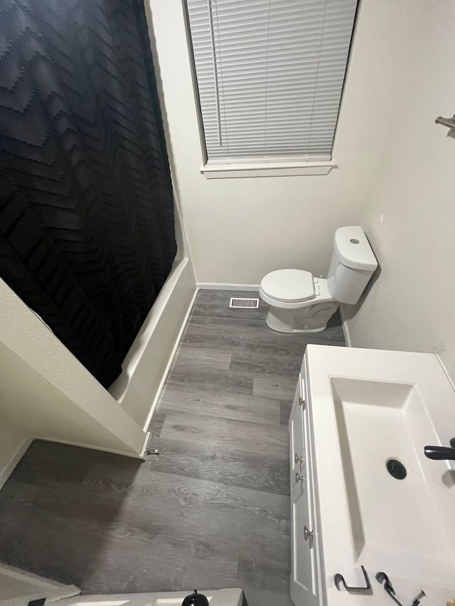 Small bathroom with toilet, sink, shower curtain, and window blinds on gray wood-look flooring.