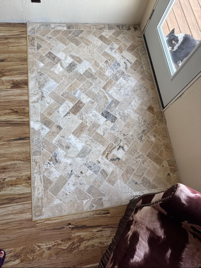 Small beige patterned rug on wood floor beside a door, with a dog’s face visible at the right edge.