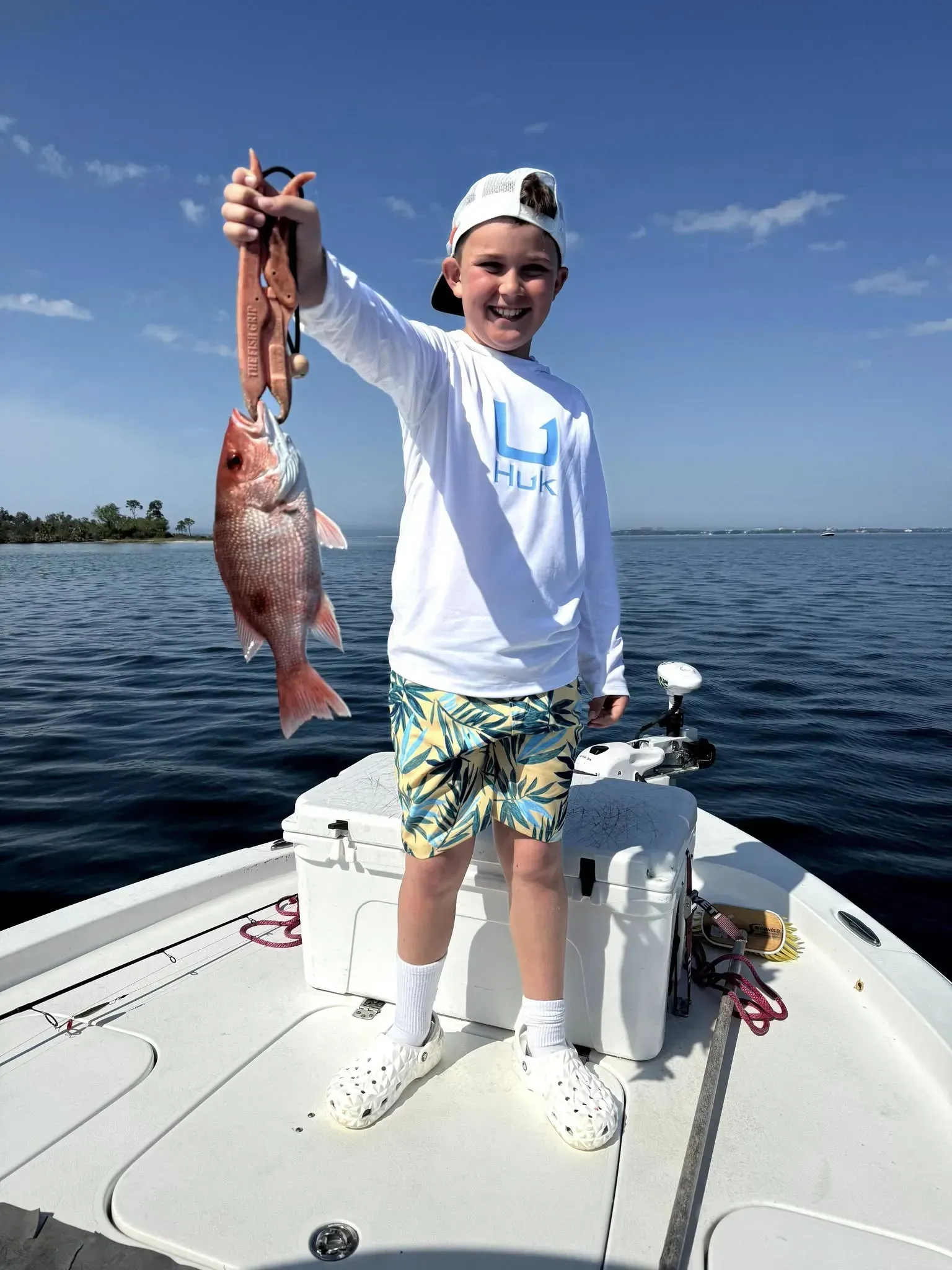 Child on a boat holding up a large caught fish over calm blue water.