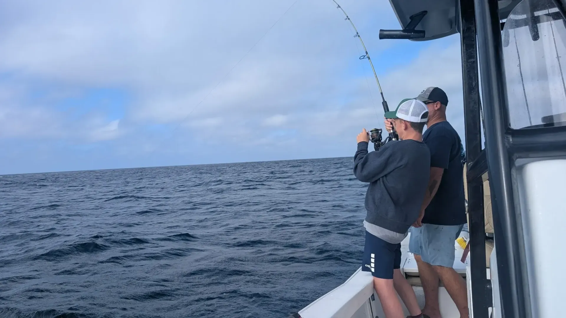 Two anglers on a boat reeling in fishing lines on a choppy ocean under a cloudy sky