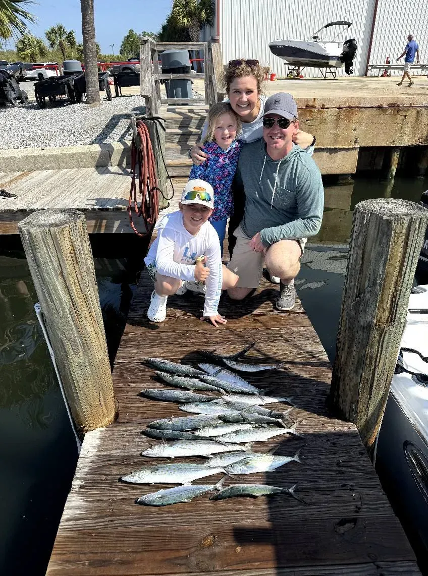 Three people crouch on a dock beside a pile of fish, with boats and water in the background.