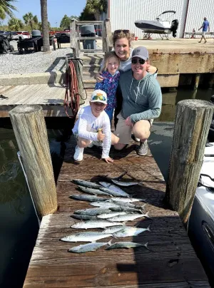 Three people kneel on a dock beside a row of fish, with boats and a harbor in the background