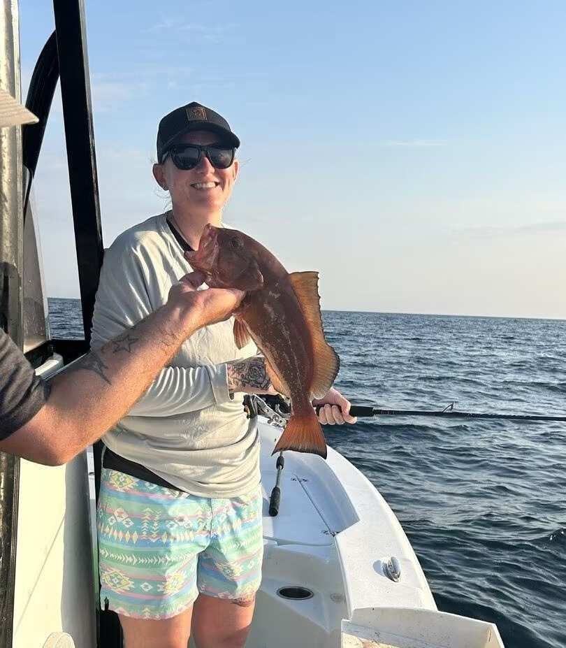 Person on a boat holding up a large fish over the ocean, wearing sunglasses and a cap.