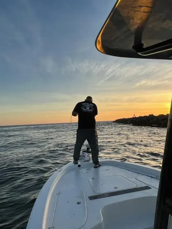 Person standing on a boat deck at sunset, fishing on calm water under a colorful sky