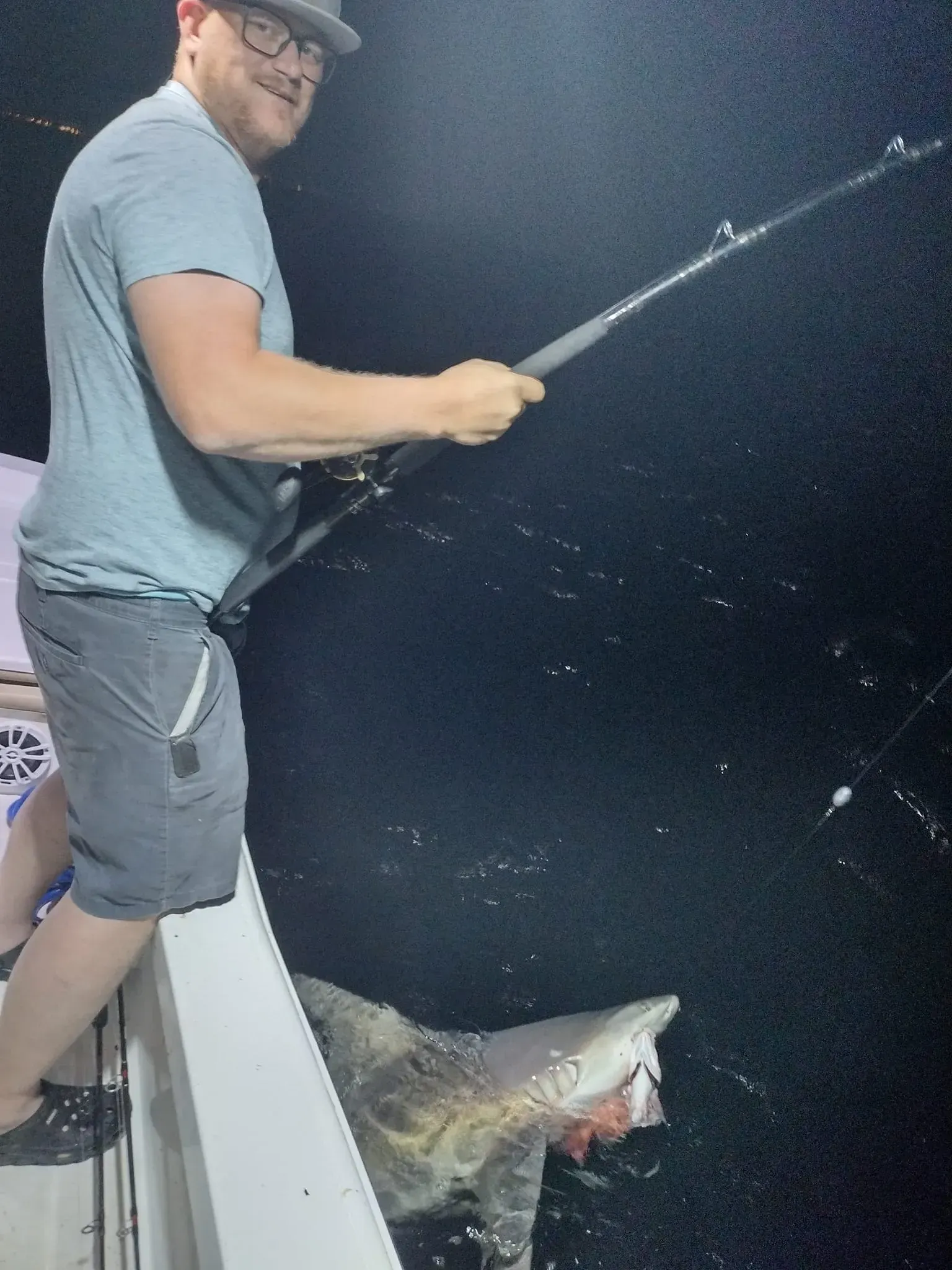 Man fishing from a boat at night, reeling in a large fish beside the hull.