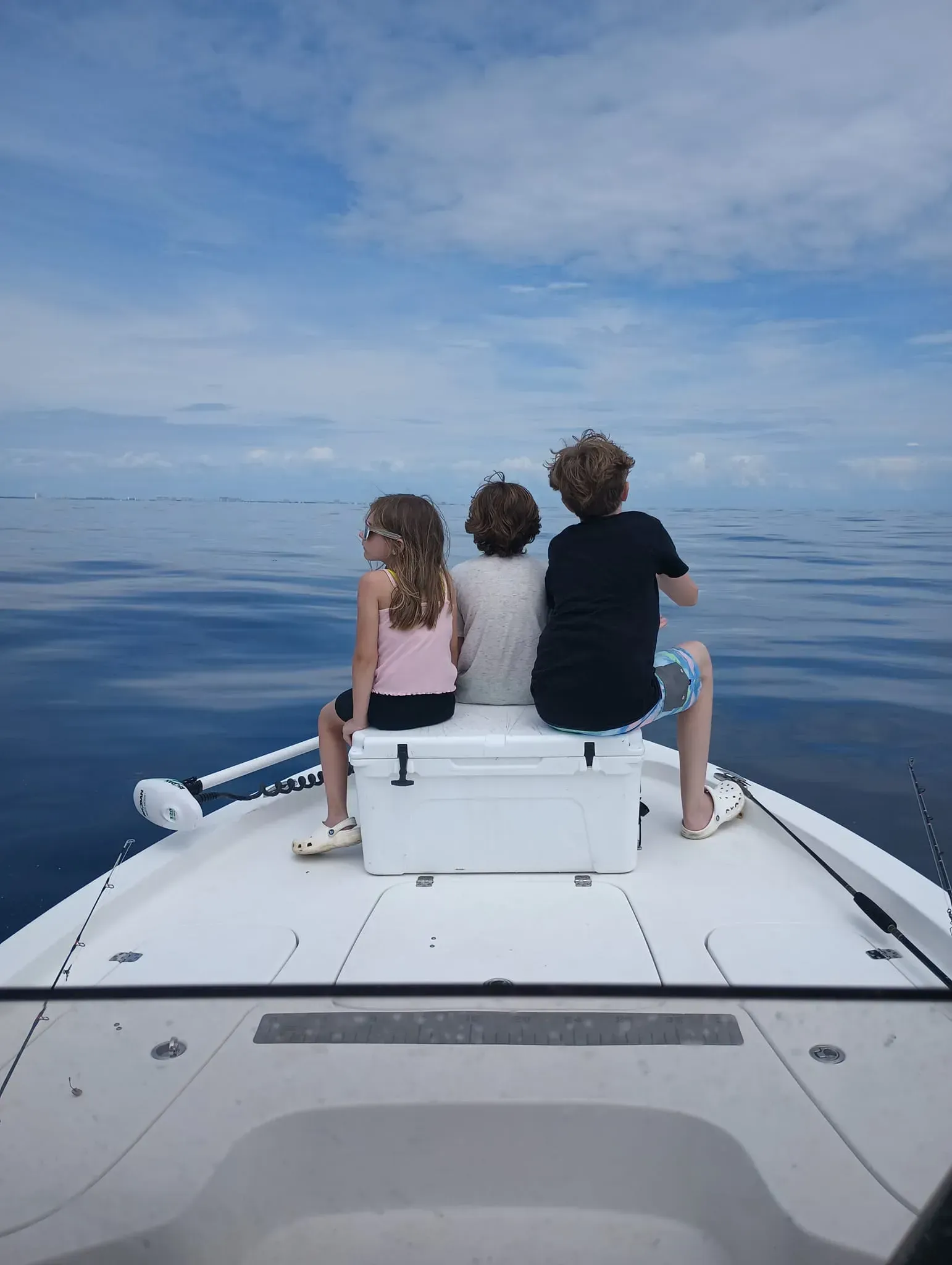 Three people sit on the bow of a boat, facing a calm open ocean under a cloudy sky.