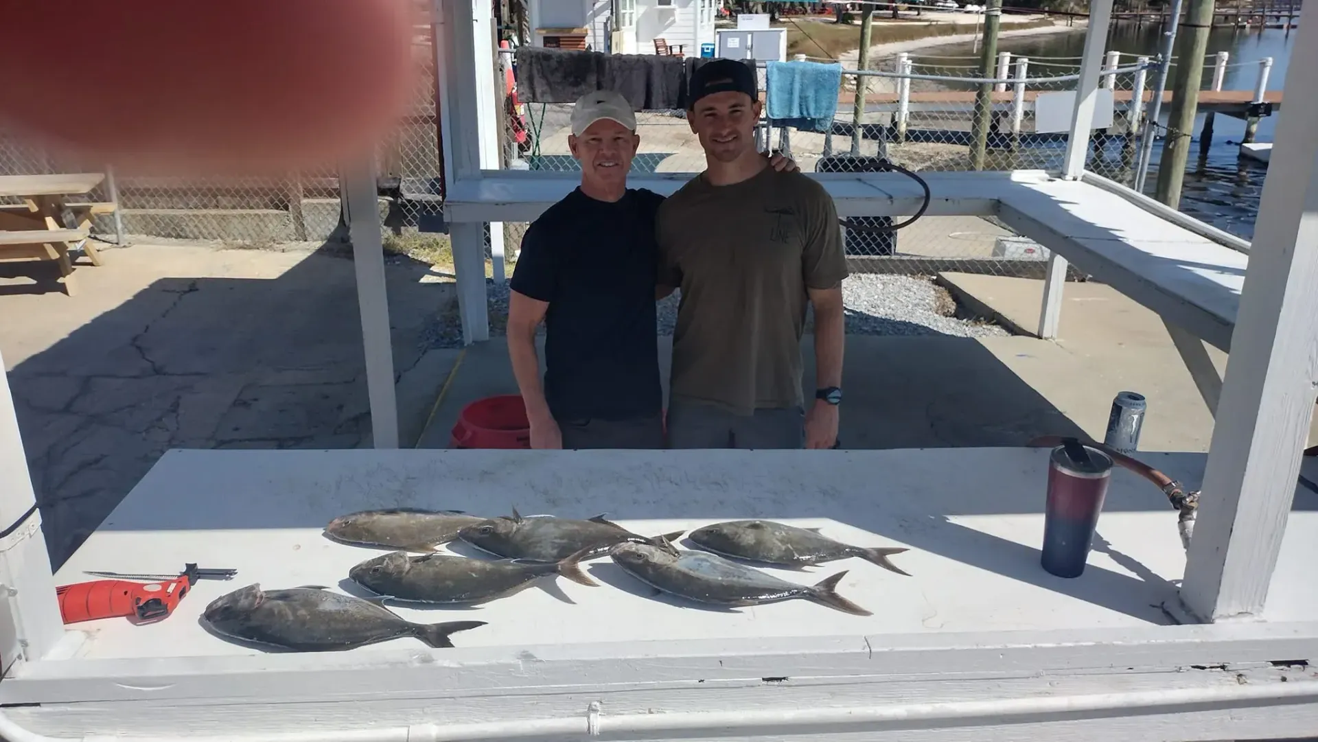 Two people stand behind a table covered with several fish under a shaded dock area.