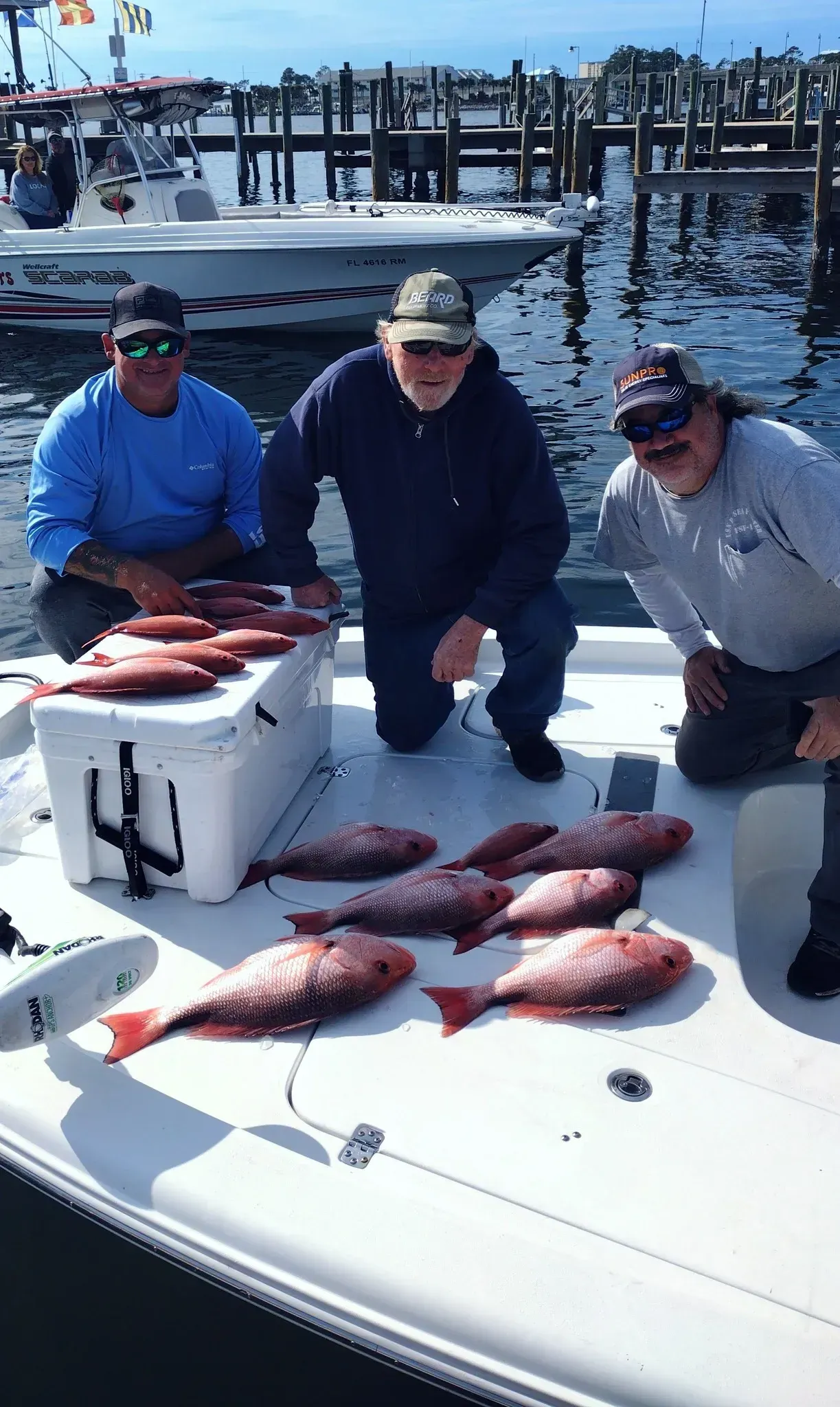 Three anglers kneel on a boat deck beside several red fish, with docks and water in the background.
