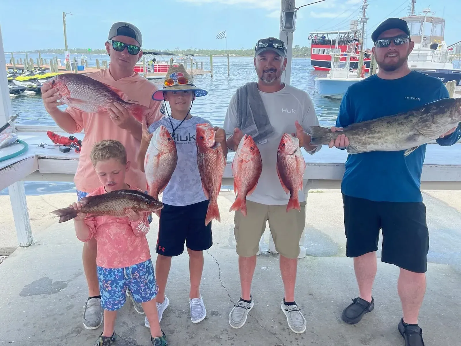 Four people on a dock holding large caught fish, with boats and water in the background.