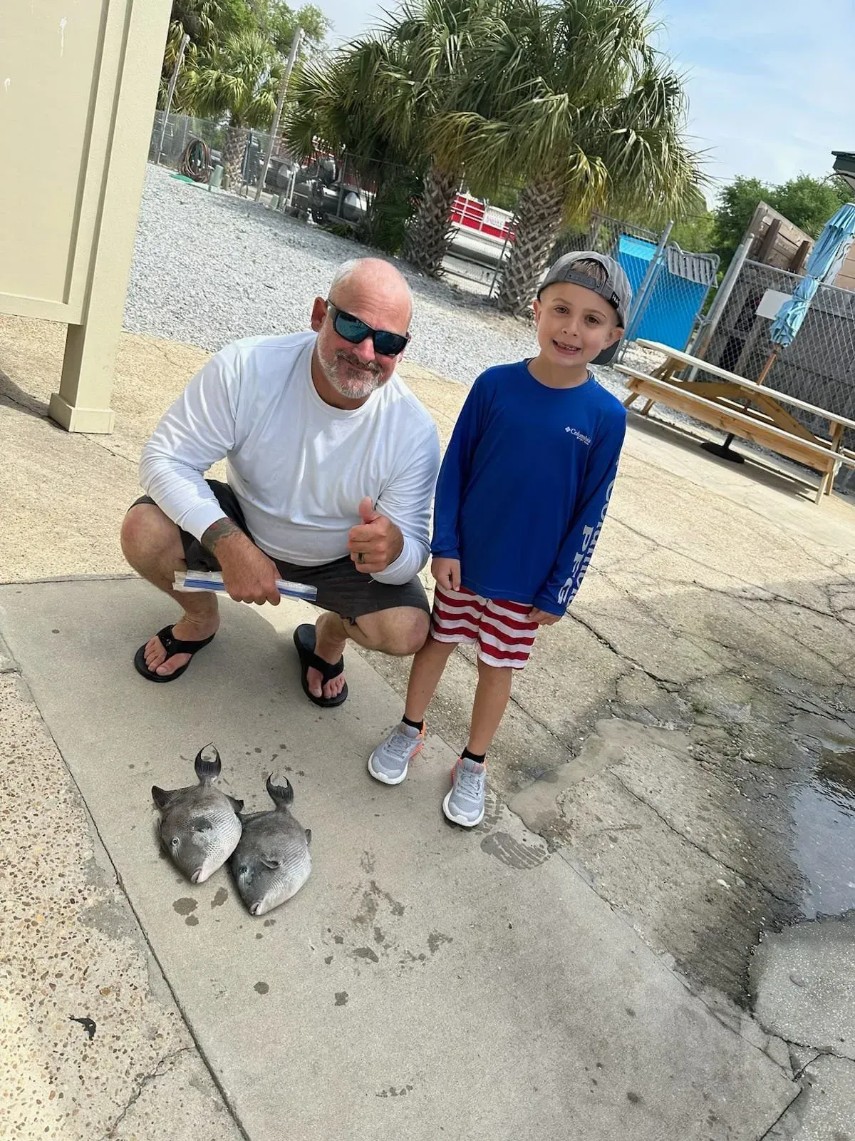Man crouches beside a child near two fish on a concrete walkway outdoors.
