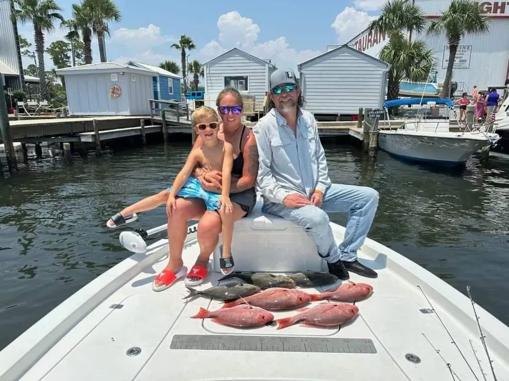 Two people on a boat with a child and several fish in the foreground, at a marina with palm trees.